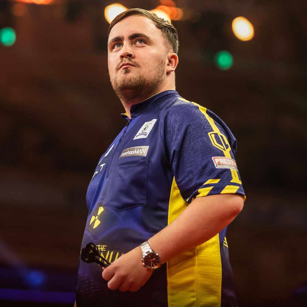 Darts player Luke Littler in a blue and yellow shirt, holding darts, looking up intently.