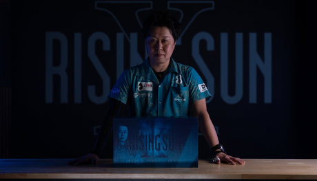 Professional darts player Haruki Muramatsu in a teal shirt, standing behind a "Rising Sun" branded box.