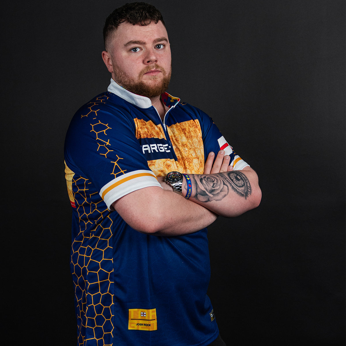 Josh Rock in a blue and gold Target darts shirt, arms crossed, against a dark background.